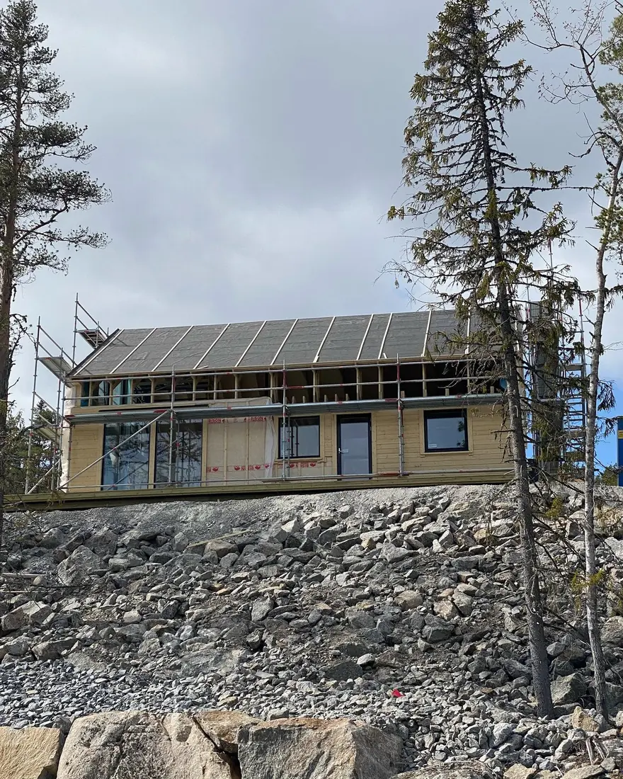 Trähus under byggnation på bergig tomt med takarbete och byggställning.