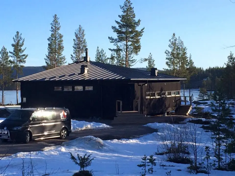Mörk träbyggnad med tak i vinterlandskap nära vatten.