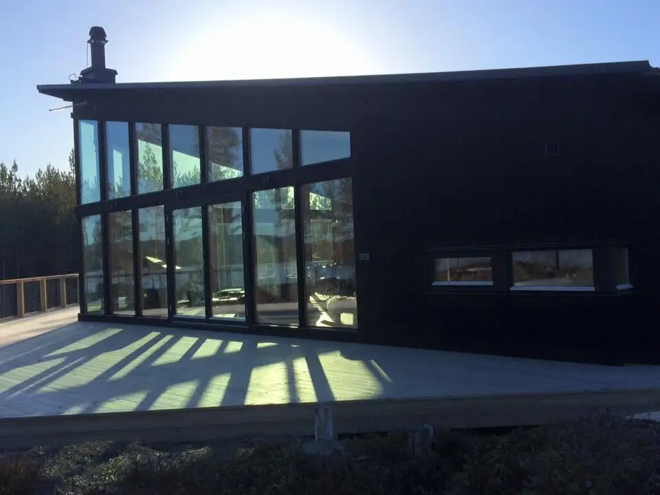Mörk träbyggnad med glasfasad och trädäck i motljus.